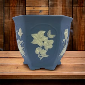 Puede incluir: Una maceta de cerámica azul con diseños florales blancos. La maceta tiene una forma hexagonal y un acabado liso y esmaltado.