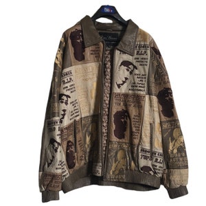 May include: A brown leather jacket with a zipper closure. The jacket is covered in a collage of newspaper clippings and images featuring the rapper Tupac Shakur. The text on the jacket includes "R.I.P." and "Tupac's Last Words."