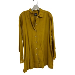 May include: A mustard yellow long-sleeve button-down shirt with a collar and a front placket featuring light-colored buttons. The shirt has a Gap brand label at the inner neckline and is displayed on a gray plastic hanger against a white background.
