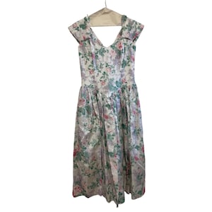 May include: A vintage floral print dress with a V-neckline and cap sleeves. The dress features a white background with pink, green, and purple floral patterns. It hangs on a wooden hanger.