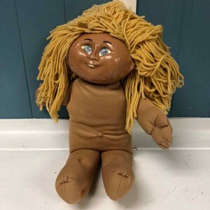 Puede incluir: Una muñeca vintage con cara y cuerpo marrones, y cabello de hilo amarillo claro. La muñeca tiene ojos azules y viste un atuendo marrón. La muñeca está sentada.