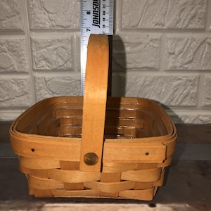 May include: A small, square woven basket with a wooden handle. The basket is made of light brown woven material and features a handle attached with a metal rivet. The basket measures approximately 18 cm in length and width.