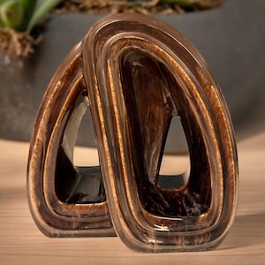 Könnte beinhalten: Zwei abstrakte braune Skulpturen mit glänzendem Finish. Die Skulpturen haben ein schlaufenförmiges, offenes Design mit einem geschichteten Aussehen. Die Farbe variiert von hell- bis dunkelbraun, mit Marmoreffekt. Die Objekte werden auf einer Holzoberfläche ausgestellt.