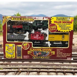 May include: A boxed G-gauge Grand Canyon Express radio-controlled train set. The set includes a red and green locomotive, various train cars, and a remote control. The box features text such as "Transmitter Controlled Train Sounds" and "40 Piece Train Set".