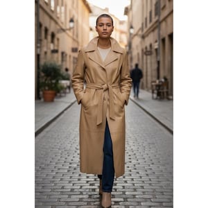 Puede incluir: Un abrigo largo de cuero color beige con cinturón en la cintura y solapas grandes. El abrigo largo se lleva sobre una camiseta color crema y vaqueros azul oscuro. La modelo camina por una calle adoquinada.