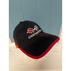 Może przedstawiać: Czarna czapka z daszkiem z czerwonym rondem i akcentami. Czapka zawiera napis "Corvette" w białym skrypcie, wraz z logo skrzyżowanych flag. Z boku czapki znajduje się płomień.