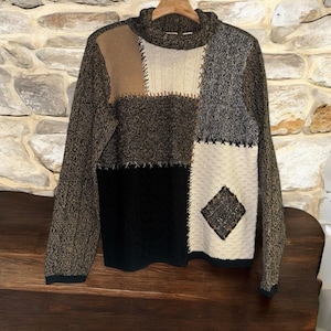 Könnte beinhalten: Ein Patchwork-Pullover mit hohem Kragen. Der Pullover hat verschiedene strukturierte Bahnen in Braun-, Schwarz-, Grau- und Cremetönen, die mit kontrastierendem Faden zusammengenäht sind. Auf der Vorderseite ist ein rautenförmiges Detail zu sehen.