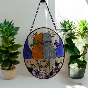 Puede incluir: Una obra de arte circular de vidriera que representa dos gatos, uno naranja y otro gris, sobre un fondo de luz de luna. Mariposas y flores adornan la parte inferior. La pieza está suspendida por una cadena negra.