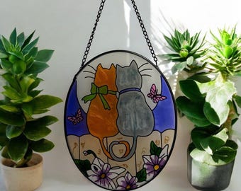 Puede incluir: Una obra de arte circular de vidriera que representa dos gatos, uno naranja y otro gris, sobre un fondo de luz de luna. Mariposas y flores adornan la parte inferior. La pieza está suspendida por una cadena negra.