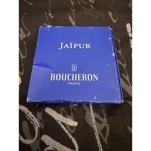 May include: A rectangular blue box with white text that reads JAÏPUR, a small logo, and the text BOUCHERON PARIS. The box has visible wear and white scuff marks along the edges and corners, indicating previous use and handling of the container.