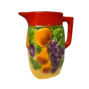 May include: A ceramic pitcher with a red rim and handle, and a yellow base. The pitcher is decorated with a colorful fruit design, including oranges, pears, grapes, and plums, set against a green leafy background.