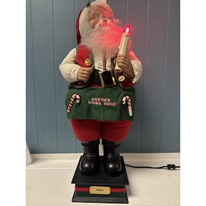 Puede incluir: Una figura de Papá Noel con chaleco y pantalón rojos, barba blanca y gafas. Sostiene una vela con una llama roja. El delantal dice "SANTA'S WORK SHOP". La figura se encuentra sobre una base negra con la palabra "NOEL".