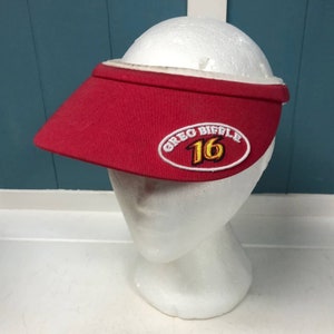 May include: A red visor with a white interior and trim. The visor features an embroidered oval patch with the text "GREG BIEFLE 16" in white and gold. It is displayed on a white mannequin head.