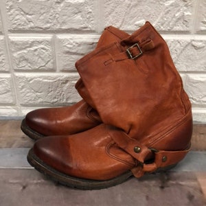 Könnte beinhalten: Braune Lederstiefel mit Knopfschließe und einem Riemen mit einer Metallschnalle um den Knöchel. Die Stiefel haben einen Used-Look und einen dicken Absatz.