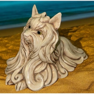 Puede incluir: Figura de perro de cerámica color crema con pelo detallado y ojos expresivos. El perro está tumbado, con la cabeza y las patas delanteras visibles. La figura tiene un acabado brillante y está sobre un fondo borroso de arena y agua.