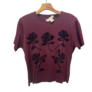 May include: A burgundy short-sleeved shirt with a crew neck, adorned with five black rose designs. The shirt is hanging on a wooden hanger. The fabric has a textured appearance.