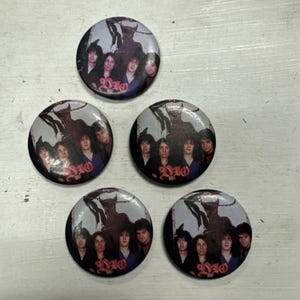 Puede incluir: Cinco botones redondos, negros y blancos con una imagen de la banda de rock Dio, con una figura de diablo en el fondo. Los miembros de la banda visten ropa oscura y el texto "DIO" está impreso en rosa en cada botón.