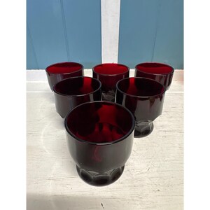 Puede incluir: Un conjunto de cinco vasos de vidrio rojo rubí a juego dispuestos en grupo. Cada vaso presenta una forma de cuenco lisa y redondeada que se estrecha hacia una base hexagonal facetada, creando un perfil consistente en los cinco recipientes.