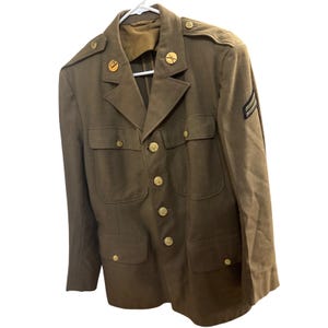 May include: Olive drab military jacket with gold-coloured buttons and insignia. The jacket has four front pockets and a sergeant's stripes patch on the sleeve. The jacket is hanging on a white hanger.