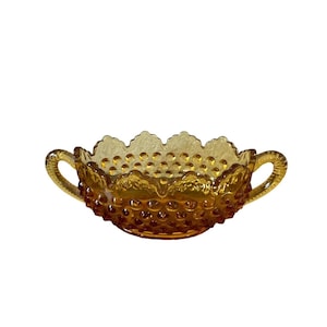 Puede incluir: Cuenco de vidrio color ámbar con dos asas. El cuenco tiene una superficie texturizada con pequeñas protuberancias redondeadas y un borde festoneado. Las asas son curvas y tienen un diseño texturizado. Este cuenco vintage es una pieza decorativa.