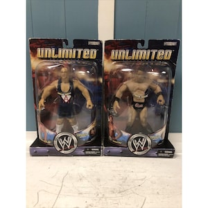 Puede incluir: Dos figuras de acción de luchadores en embalaje de plástico. Las figuras visten un atuendo azul y otro con calzoncillos negros. El embalaje dice "UNLIMITED" y presenta el logotipo de la WWE.