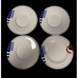 May include: Four white ceramic plates with a geometric design. Each plate features a blue, grey, and black rectangular pattern, with a thin black line around the rim. One plate has additional red, green, and yellow accents.