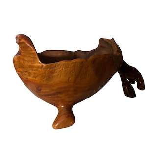 May include: A polished wooden bowl with a natural, irregular live edge rim and a deep, rounded basin. The bowl features a warm, light brown wood grain pattern and is supported by a single, thick, integrated wooden foot at the base.