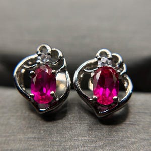 May include: A pair of silver-toned stud earrings featuring a central oval-cut vibrant pink gemstone surrounded by a curved metal frame and a small clear accent stone at the top of each piece, showcasing a polished metallic finish and faceted gems.
