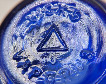 May include: Close-up of the bottom of a cobalt blue glass bottle. The base features embossed text and a triangle symbol. The glass has a textured surface, and the details are highlighted by the light.