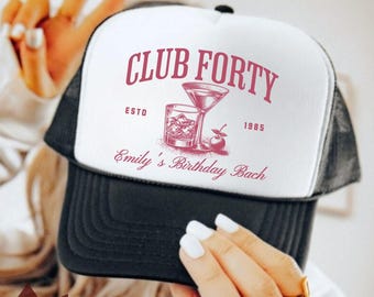 May include: A black and white trucker hat with the text "CLUB FORTY" in pink, along with "ESTO 1985" and "Emily's Birthday Bach". The hat features a cocktail and a glass of whiskey graphic.