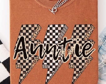 May include: Auntie t-shirt in a burnt orange color. The word "Auntie" is printed in a black script font, with a checkered and leopard print lightning bolt design. The shirt is folded and has a silver chain necklace.