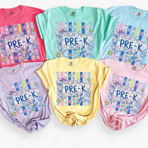 May include: Six short-sleeved crew neck t-shirts in pink, mint green, light blue, lavender, yellow, and light pink. Each shirt features a central graphic of a pennant flag with the text PRE-K, surrounded by blue bows, stars, and stripes.