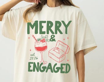 Puede incluir: Una camiseta de manga corta color blanco roto con el texto verde MERRY & ENGAGED. El gráfico incluye una copa de cóctel roja con un bastón de caramelo y acebo, un joyero rojo abierto con un anillo y el texto est 2026 en letras verdes.