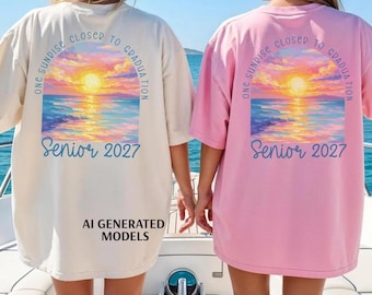 May include: Two oversized t-shirts, one cream and one light pink, featuring a back graphic of a sunset over the ocean. The text reads "ONE SUNRISE CLOSER TO GRADUATION" in an arc above the sun and "Senior 2027" in blue script below the water.