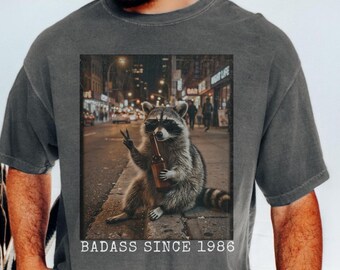 May include: Dark grey t-shirt featuring a graphic of a raccoon in sunglasses holding a bottle, with the text "BADASS SINCE 1986". The raccoon is sitting on a city street at night.