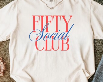 May include: Cream-colored t-shirt with the words "FIFTY Social CLUB" in red and blue lettering. The shirt is laid flat, with a silver necklace at the neckline. Other items include white sneakers, a brown hat, and a pair of socks.
