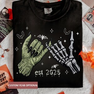 May include: Black t-shirt featuring a green zombie hand and a white skeleton hand, both pinky-swearing. The design includes bats, stars, and the text "est 2025". The phrase "CUSTOM YEAR OPTIONAL" is also visible.