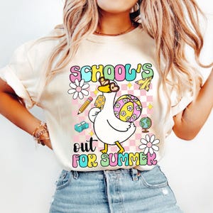 Pu&ograve; includere: T-shirt color crema con una grafica colorata che recita "SCHOOL'S OUT FOR SUMMER". La grafica presenta un'oca dei cartoni animati con occhiali da sole e che tiene in mano un pallone da spiaggia, una matita e un mappamondo. Il design include fiori e un aeroplano di carta.