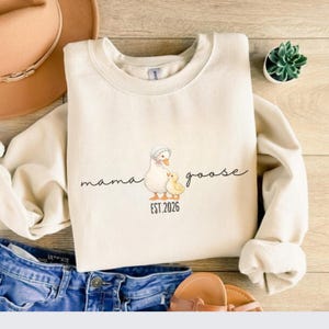 May include: Cream-colored sweatshirt with a Mama Goose graphic. The design features a mother goose and a baby chick, with the words "mama goose" and "EST. 2026" in a script font. The sweatshirt is paired with blue jeans and sandals.