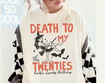 May include: Cream-colored t-shirt with the text "DEATH TO MY TWENTIES" in orange. A witch illustration is in a martini glass. The shirt also includes "est 1996" and "Emily's Spooky Birthday". The person is wearing a black and white checkered cardigan.