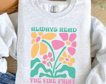 Puede incluir: Sudadera gris claro con un diseño floral en rosa, amarillo y turquesa. El texto "ALWAYS READ THE FINE PRINT I'M PREGNANT" está impreso encima y debajo del gráfico floral. La sudadera está hecha de un material suave.