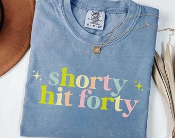 May include: A blue Comfort Colors t-shirt with the phrase "shorty hit forty" in colorful text. A gold necklace with a pendant rests on the shirt. A brown hat is partially visible in the upper left corner.