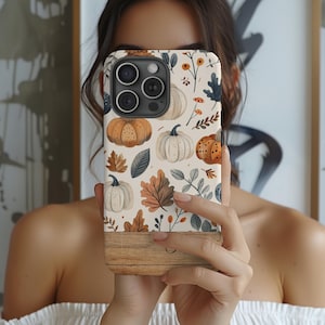 May include: A phone case featuring a fall-themed design with pumpkins, leaves, and floral elements in shades of orange, brown, and cream. The bottom portion of the case has a wood-grain texture.