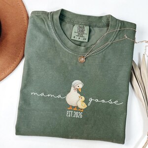 May include: Olive green Comfort Colors t-shirt with a "mama goose" graphic and "EST. 2026" text. The graphic features a goose and gosling in a vintage style. A silver necklace with a pendant is also visible.