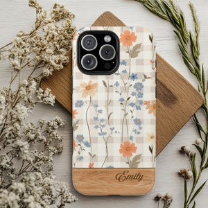 May include: A floral phone case featuring a beige plaid pattern with orange and blue flowers. The bottom portion is a light wood grain, with the name "Emily" engraved. The case is designed for a smartphone with multiple camera lenses.