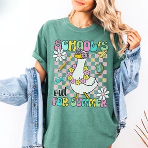 Pu&ograve; includere: T-shirt verde con la scritta "SCHOOL'S OUT FOR SUMMER" in un font retr&ograve;. La maglietta presenta un'oca dei cartoni animati con occhiali da sole e una ghirlanda di fiori, con grafica a tema scolastico.