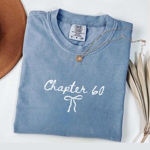 May include: A light blue Comfort Colors t-shirt with the text "Chapter 60" in white script and a bow graphic. A gold necklace with a pendant rests on the shirt. A brown hat and dried plant are in the background.