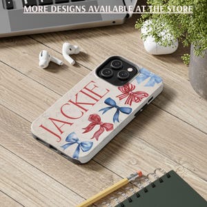 Puede incluir: Una funda de tel&eacute;fono con el nombre "JACKIE" en escritura roja, adornada con ilustraciones de lazos rojos y azules. La funda est&aacute; sobre una superficie de madera, con el texto "MORE DESIGNS AVAILABLE AT THE STORE" visible. Otros elementos incluyen auriculares, un l&aacute;piz y un cuaderno.