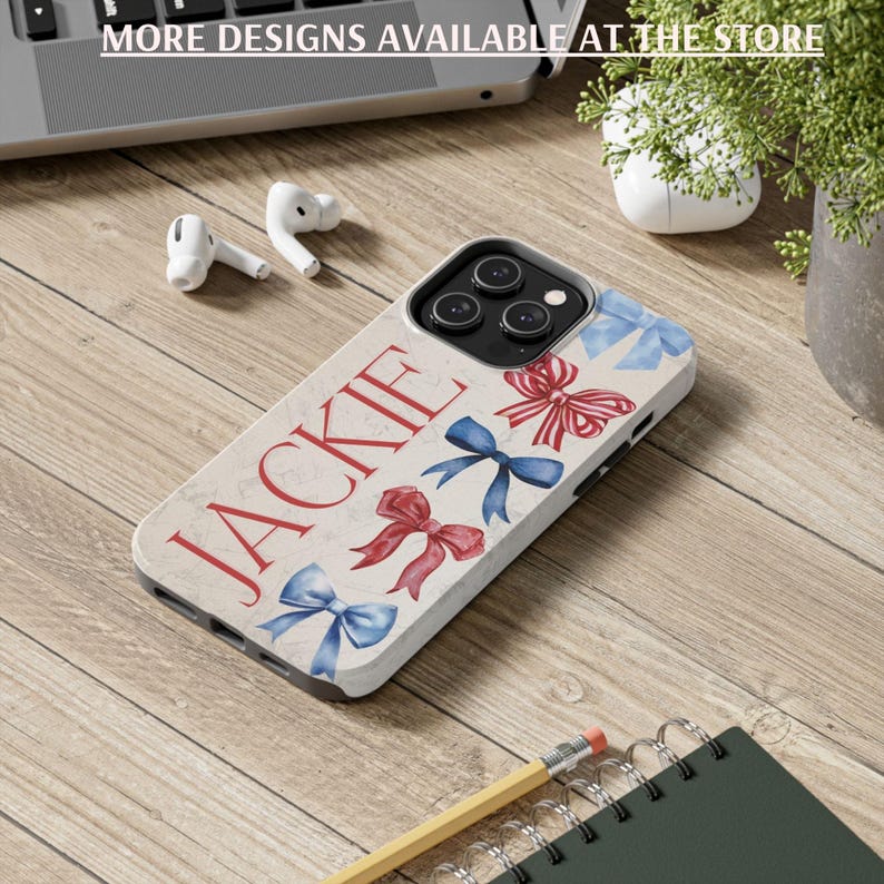Puede incluir: Una funda de tel&eacute;fono con el nombre "JACKIE" en escritura roja, adornada con ilustraciones de lazos rojos y azules. La funda est&aacute; sobre una superficie de madera, con el texto "MORE DESIGNS AVAILABLE AT THE STORE" visible. Otros elementos incluyen auriculares, un l&aacute;piz y un cuaderno.