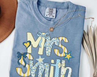 Puede incluir: Una camiseta azul de manga corta con cuello redondo y un estampado gráfico que dice "Mrs Smith" en letras grandes y estilizadas. Las letras presentan rayas amarillas, cuadros vichy azules y motivos florales, acentuados con lazos azules y estrellas amarillas.